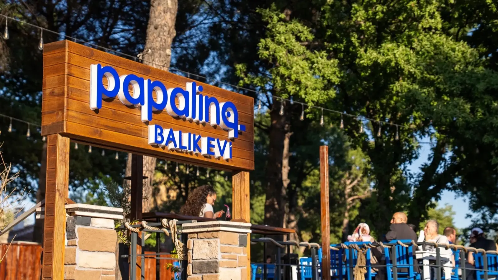 Çamların altında ahşap Papalina Balık Evi tabelası, arkada mavi sandalyeli teras